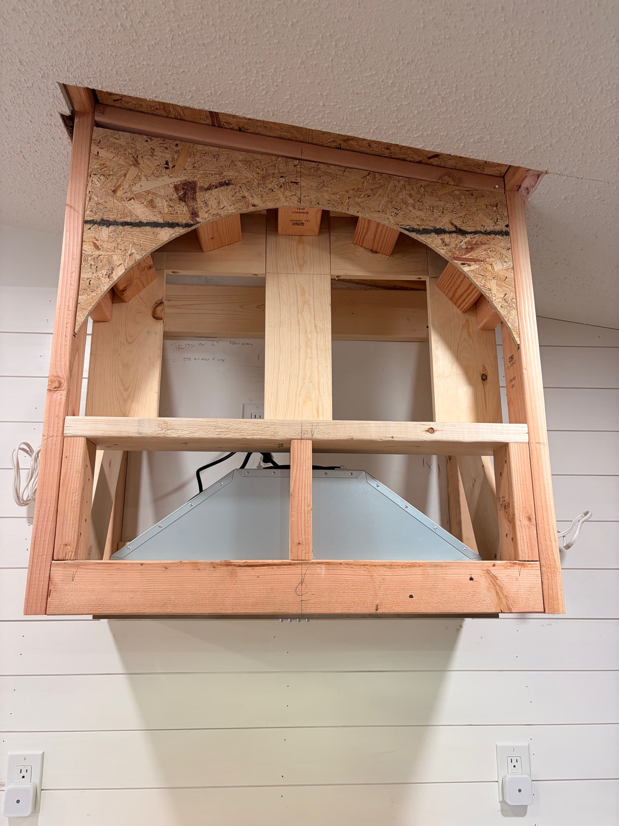 Unfinished wooden range hood frame with curved OSB panels and a metal vent insert inside.
