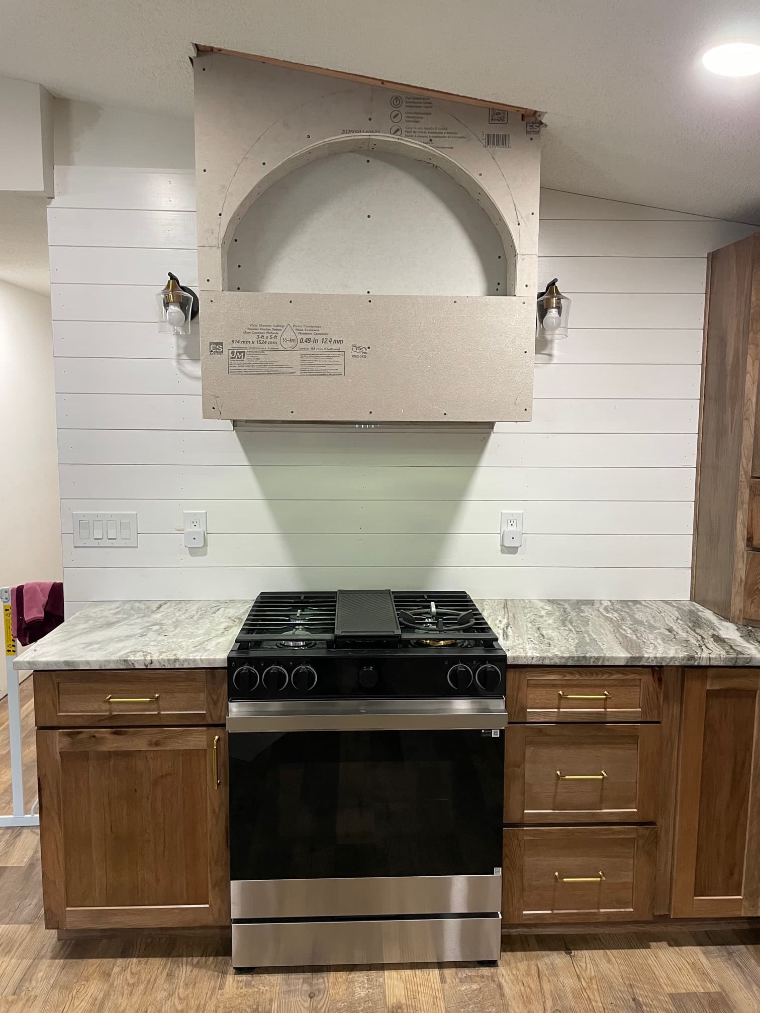 Unfinished drywall range hood above a black stove, wooden cabinets, and white shiplap wall.