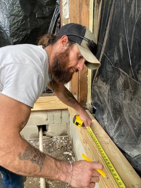 Bearded man with tattoos measuring a wooden beam using a yellow tape measure and pencil.