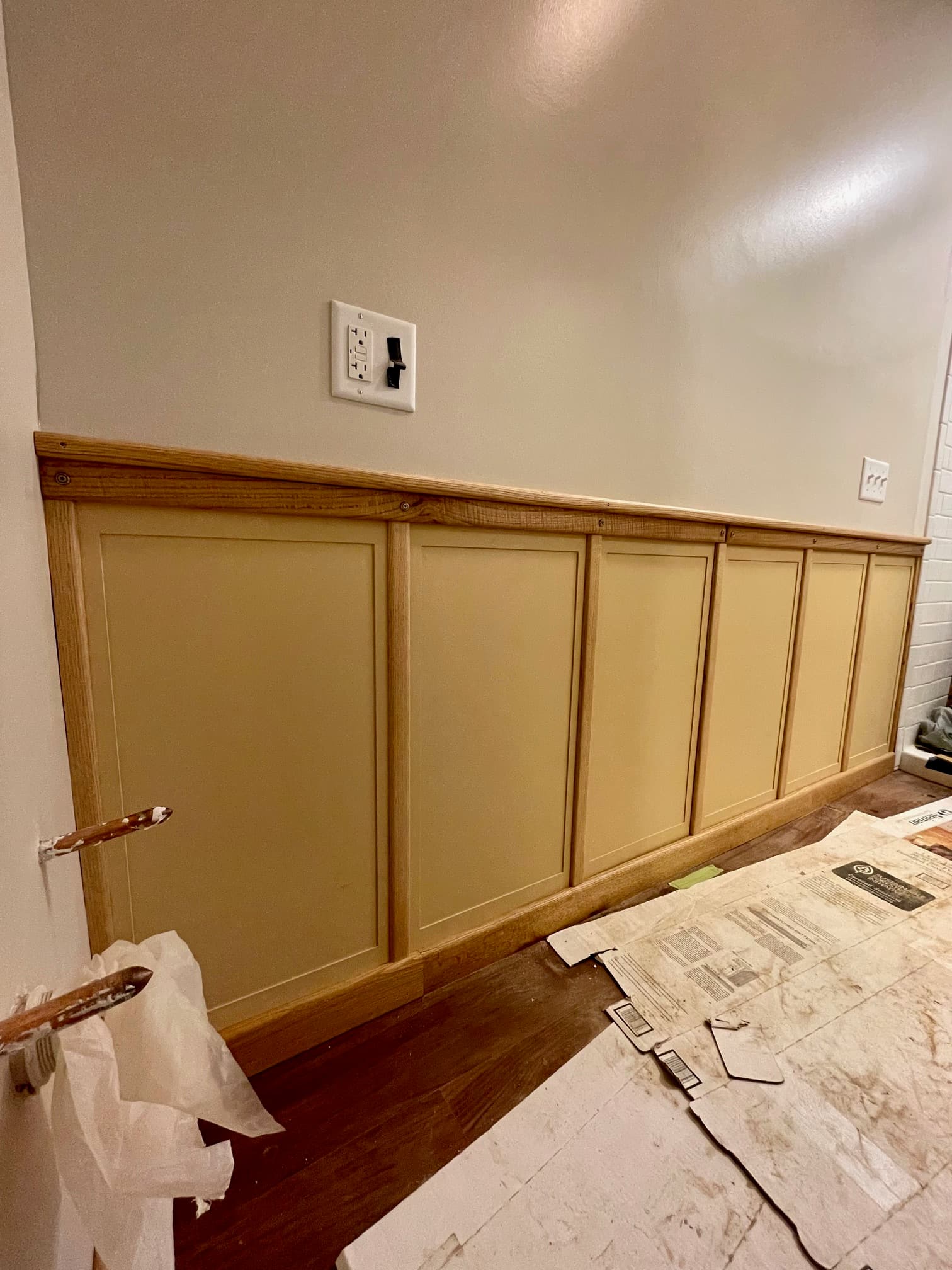 Newly installed tan wainscoting with light wood trim on a wall above dark wood floors.