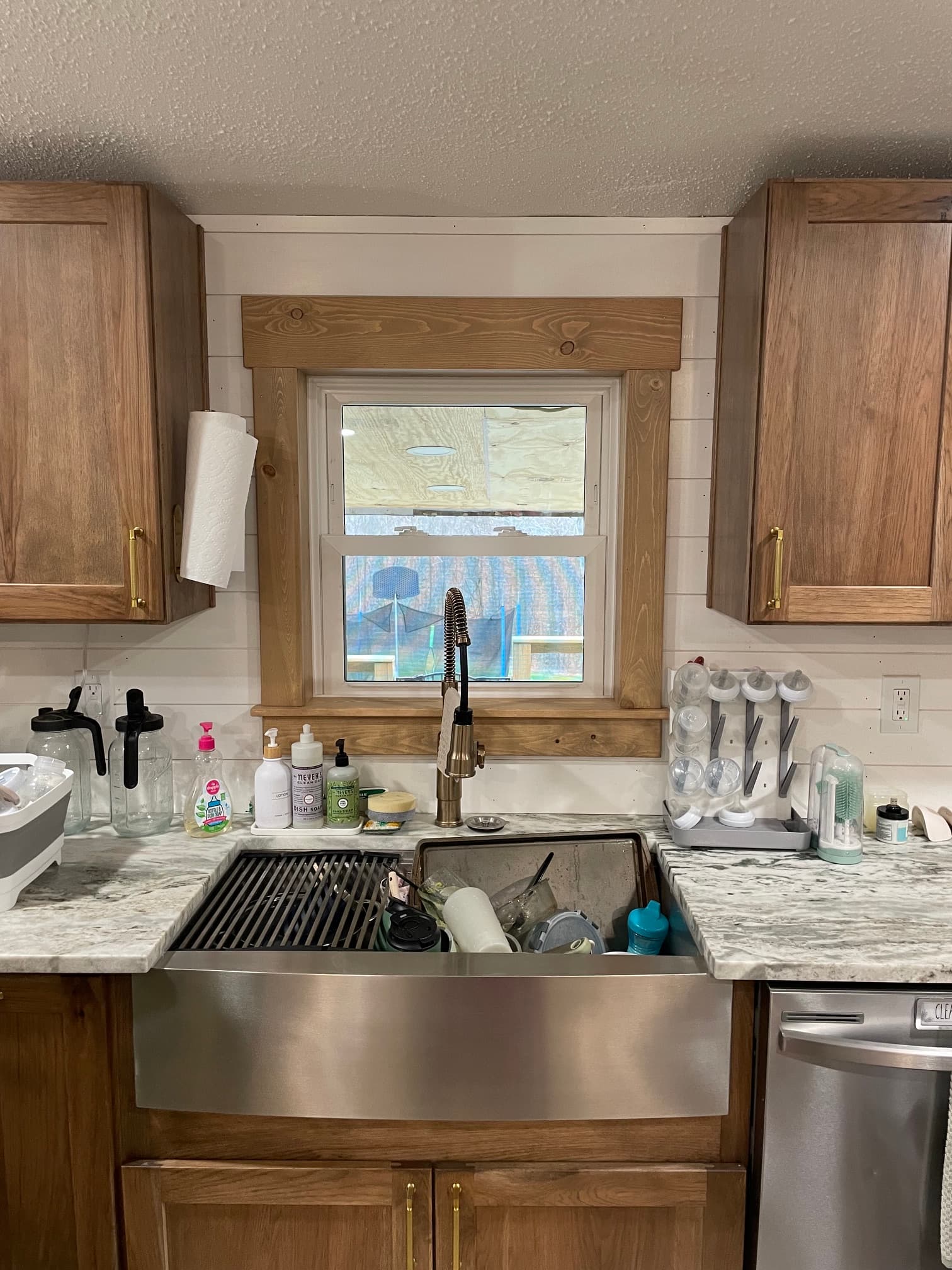 Stainless steel farmhouse sink under a window with wooden cabinets and a marble countertop.
