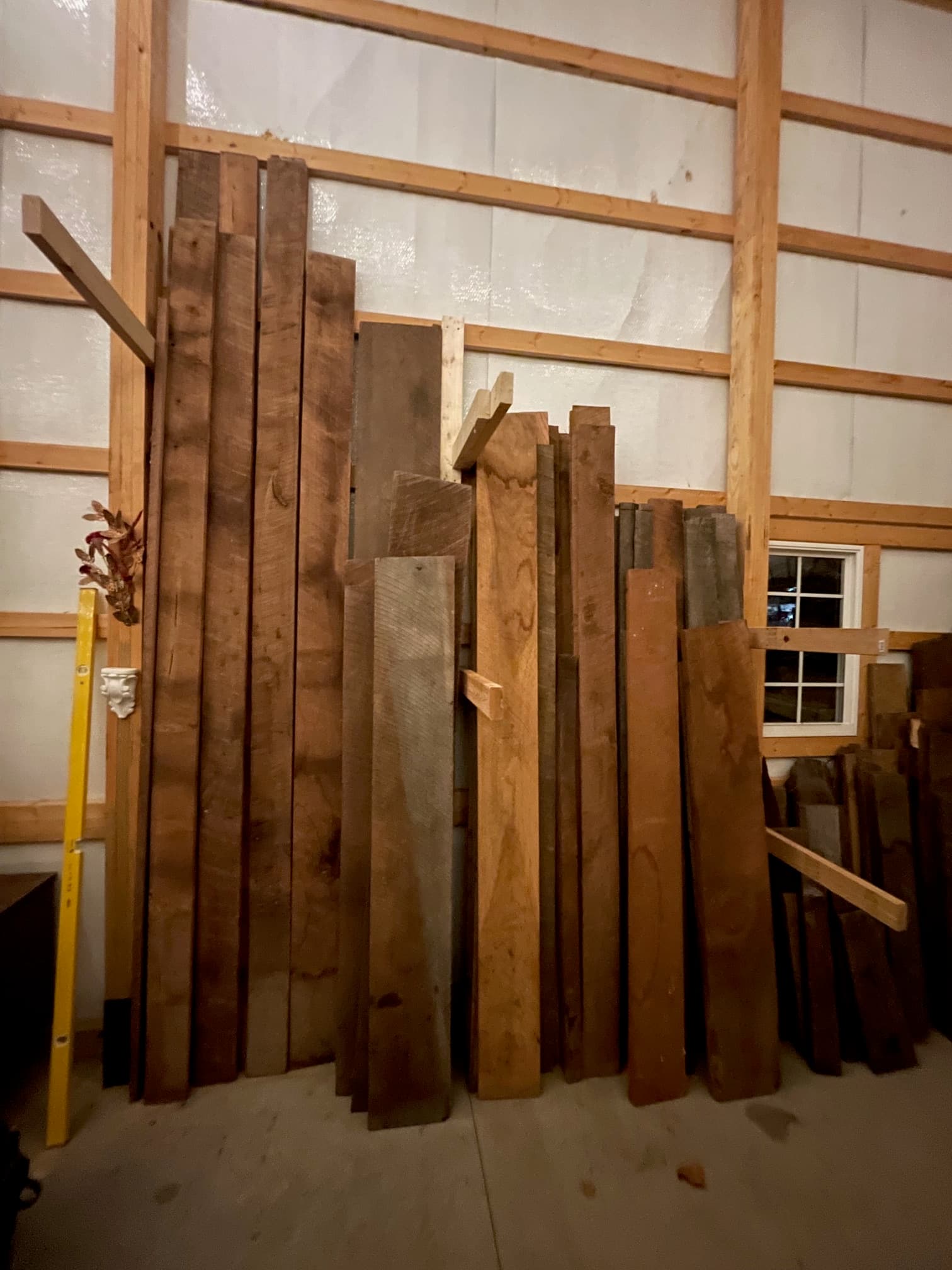 Various brown wooden planks stacked horizontally against a wall with exposed studs and a window.