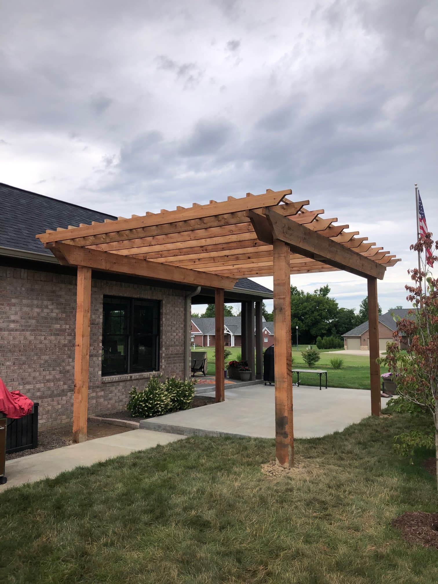 Wooden pergola structure with thick beams over a concrete patio next to a brick house.